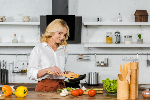 attractive middle aged woman cooking vegetables on frying pan in kitchen - Foto, Bild