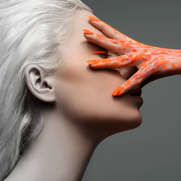 Kreatives Porträt eines schönen Mädchens mit der Hand im Gesicht, weißem Haar. die Hand mit orangefarbener Farbe bemalt - Foto, Bild