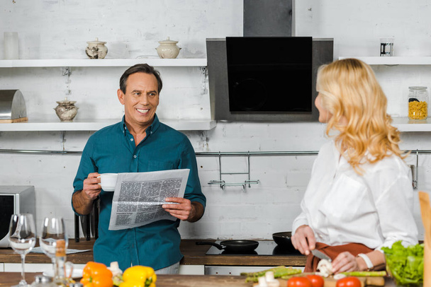 mature wife cooking and smiling husband reading newspaper in kitchen - Фото, изображение