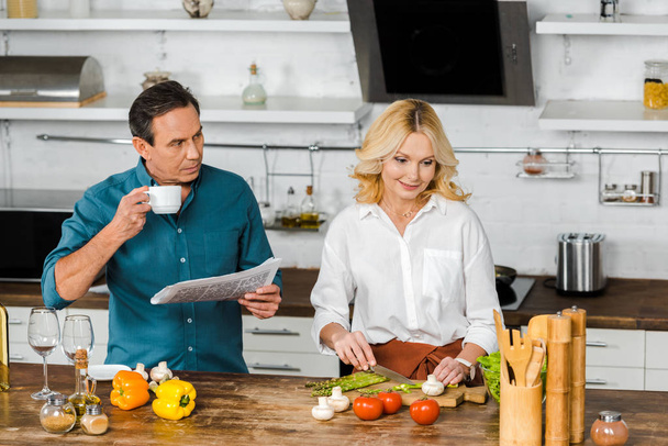 hoge hoekmening van volwassen vrouw koken en echtgenoot lezen van de krant in keuken - Foto, afbeelding