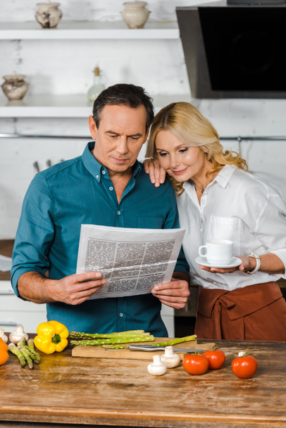 mature wife holding cup of tea and reading newspaper together with husband in kitchen - Fotoğraf, Görsel