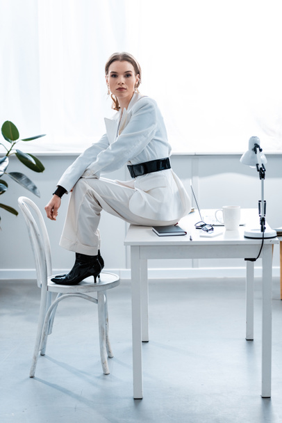 stylish businesswoman in formal wear sitting on desk and looking at camera at workplace - Fotografie, Obrázek