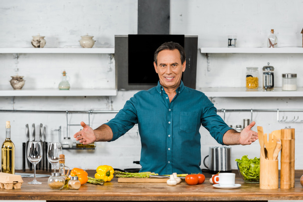 happy handsome middle aged man standing with open arms and looking at camera in kitchen - Photo, Image