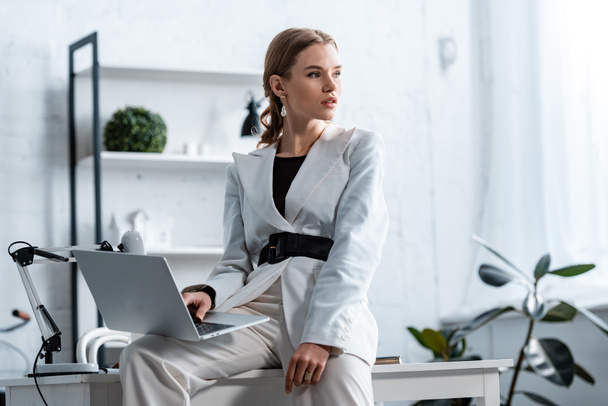 stylish businesswoman in white formal wear sitting on desk with laptop at workplace and looking away - Foto, afbeelding