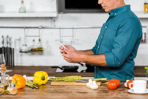 immagine ritagliata di uomo di mezza età utilizzando smartphone durante la cottura in cucina
 - Foto, immagini