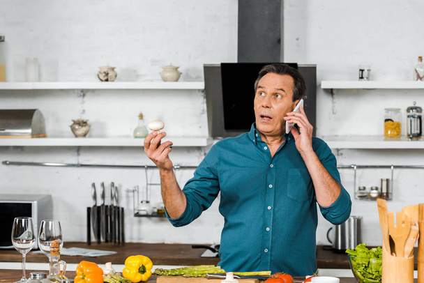 handsome middle aged man talking by smartphone while cooking in kitchen - Photo, Image