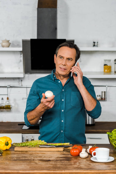 handsome middle aged man talking by smartphone and holding mushroom in kitchen - Foto, Bild