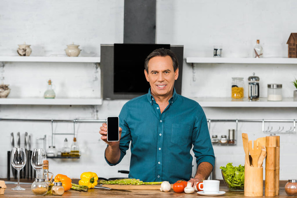lächelnder gutaussehender Mann mittleren Alters mit Smartphone mit leerem Bildschirm in der Küche - Foto, Bild
