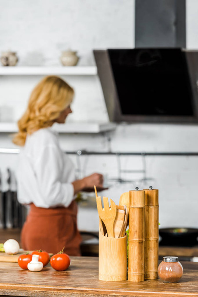 selective focus of middle aged woman holding cup of tea in kitchen, vegetables and wooden utensil on tabletop - Fotó, kép