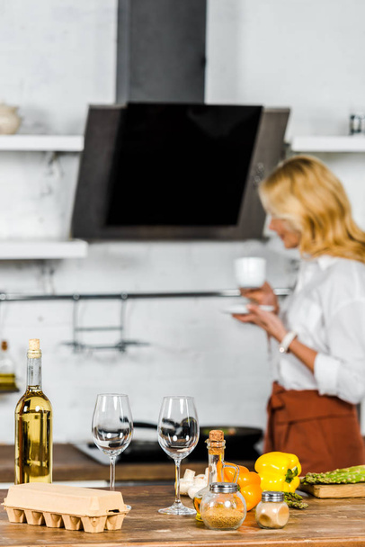 вибірковий фокус жінки середнього віку, що тримає чашку чаю на кухні, овочах та вині на столі
 - Фото, зображення
