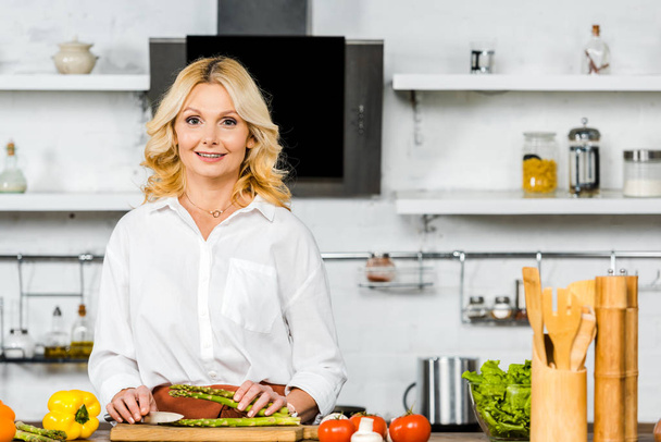 smiling beautiful middle aged woman cutting vegetables in kitchen and looking at camera - Foto, afbeelding