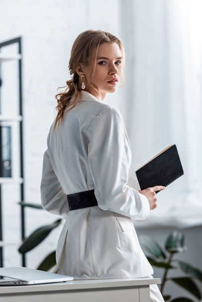beautiful businesswoman in formal wear holding notebook and looking at camera at workplace - Фото, зображення