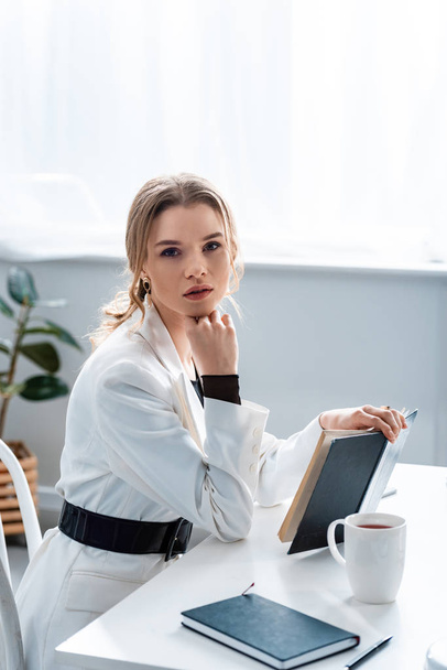 beautiful businesswoman sitting at desk, holding notebook and looking at camera at workplace - Zdjęcie, obraz