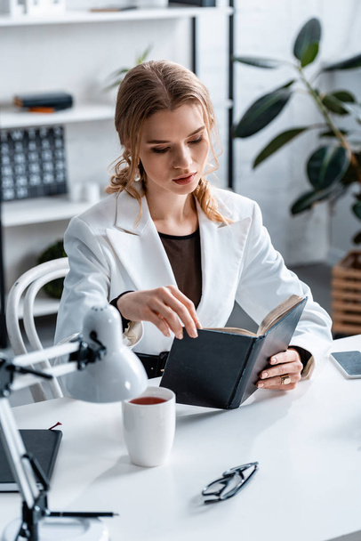 Konzentrierte Geschäftsfrau in formeller Kleidung sitzt am Schreibtisch und liest Notizbuch am Arbeitsplatz - Foto, Bild