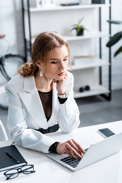 beautiful pensive businesswoman in formal wear sitting at desk and using laptop at workplace - Фото, зображення