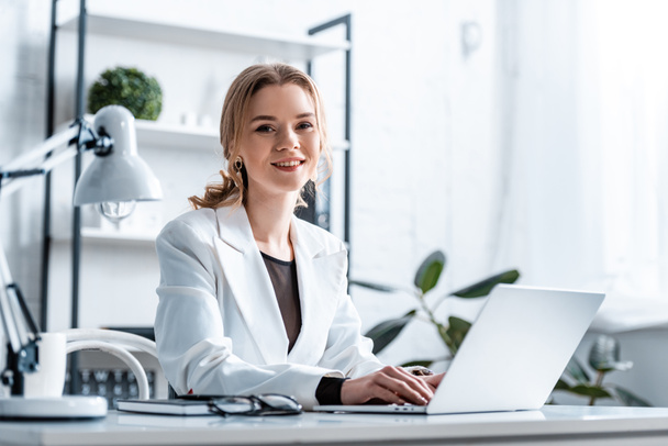 Lächelnde Geschäftsfrau in festlicher Kleidung sitzt am Schreibtisch, blickt in die Kamera und tippt am Arbeitsplatz auf den Laptop - Foto, Bild