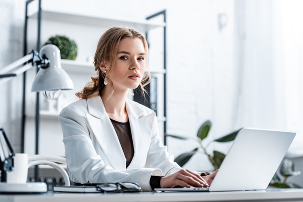 serious businesswoman in formal wear sitting at desk, looking at camera and typing on laptop at workplace - Фото, зображення