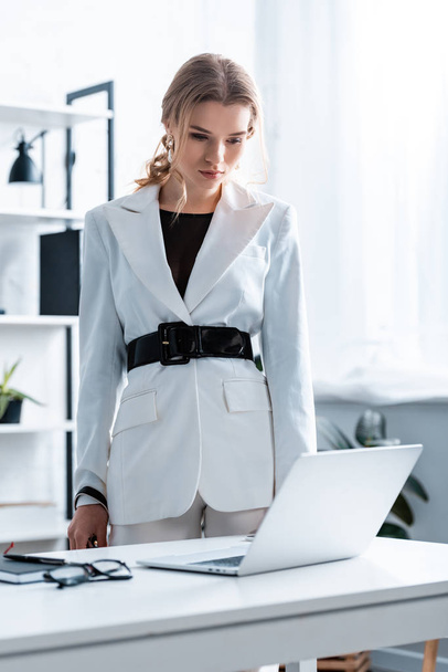 serious businesswoman in formal wear standing near desk and looking at laptop in office - Zdjęcie, obraz