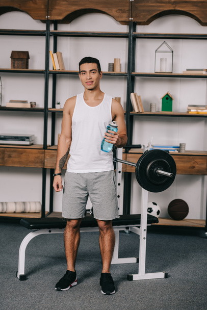 handsome bi-racial man holding sport bottle and standing near barbell  - Photo, image