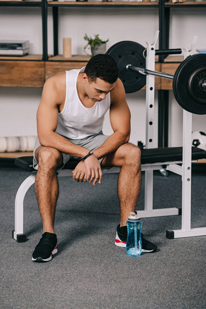 mixed race man sitting in sportswear and looking at sport bottle  - Photo, Image