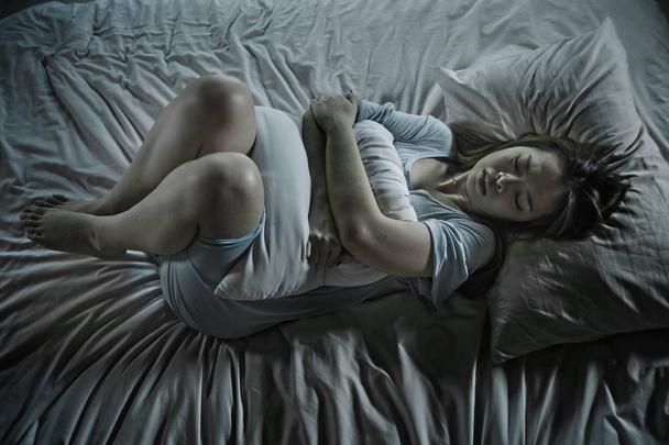 λυπημένος και καταθλιπτικός όμορφη Ασιατική Κινέζικη κοπέλα πάσχουν έμμηνος ρύση και πόνους περιόδου, αίσθημα αδιαθεσίας στο κρεβάτι τη νύχτα με πόνο στο στομάχι και κράμπες ως γυναίκα στην επώδυνη έμμηνο κύκλο, έννοια - Φωτογραφία, εικόνα