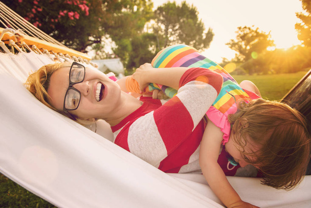 Happy mother and her little daughter enjoying free time using tablet computer while relaxing in a hammock during sunny day on holiday home garden - Фото, изображение