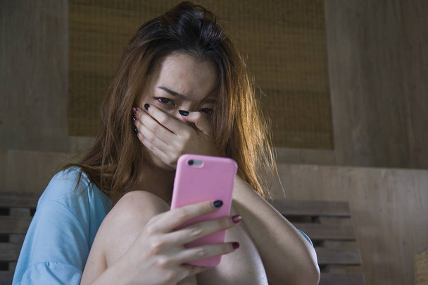 young depressed Asian Korean girl using mobile phone crying on bed at night feeling sad and depressed victim of cyber bullying or broken heart suffering depression in desperate woman expression - Foto, immagini