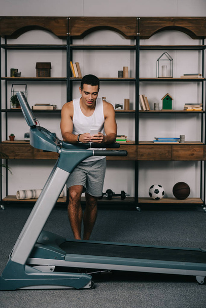 strong mixed race man using smartphone near treadmill  - Photo, Image