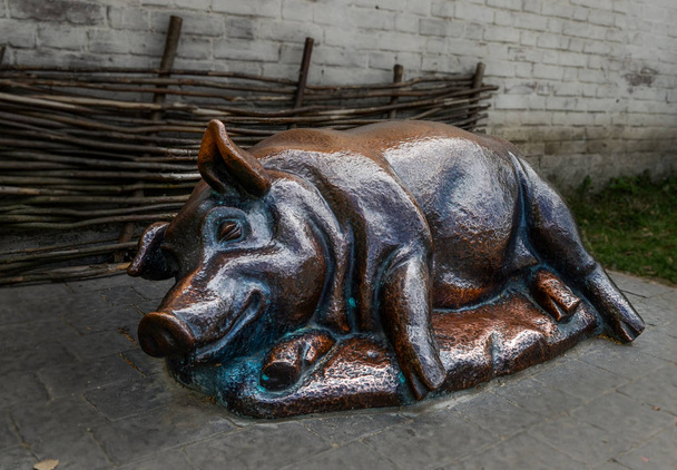 Бронзовая свинья с довольной мордой лежит на боку у забора.
 - Фото, изображение