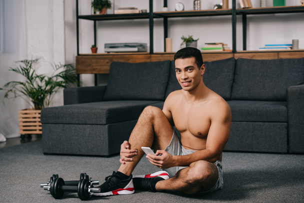 cheerful bi-racial man sitting on floor near dumbbells while using smartphone in living room  - Photo, image