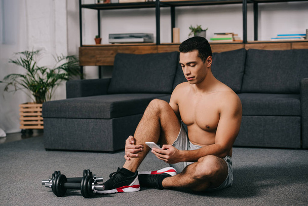 bi-faji ember rövid idő használ smartphone nappaliban padló közelében súlyzók ül  - Fotó, kép