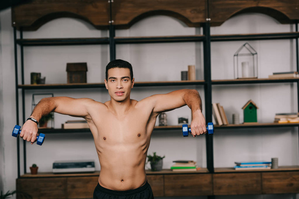 handsome bi-racial man exercising with blue dumbbells in home gym  - Photo, Image