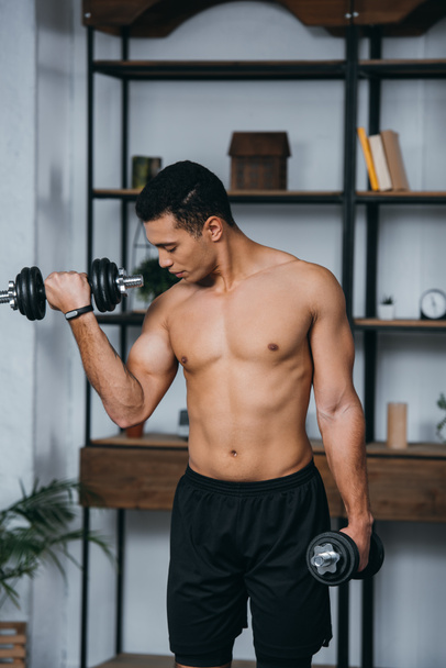 bi-racial man looking at muscles while exercising with heavy dumbbells in home gym  - Fotó, kép