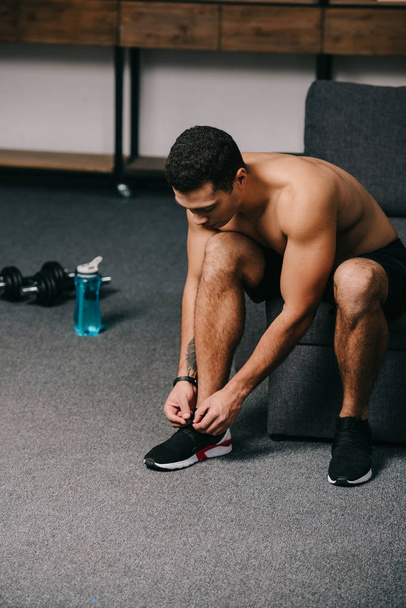 strong bi-racial man tying shoelaces near dumbbells and sport bottle in living room  - Foto, afbeelding