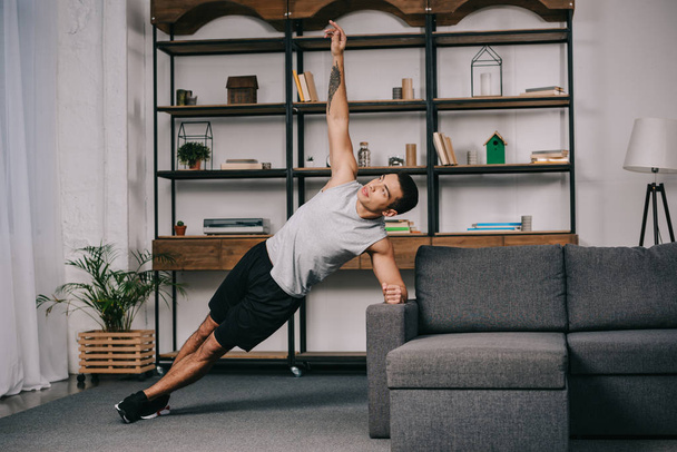 schöner gemischter Rassenmann mit Tätowierung macht Plankenübung in der Nähe des Sofas im Wohnzimmer  - Foto, Bild