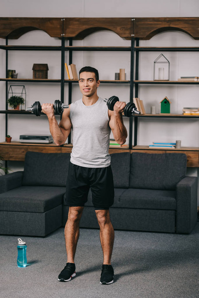 cheerful mixed race man workout with dumbbells in living room  - Photo, Image