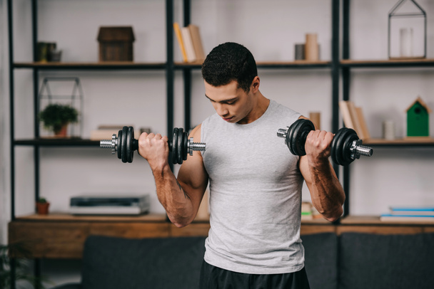 strong mixed race man training with dumbbells in living room  - Photo, Image