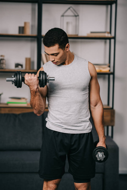 handsome mixed race man looking at muscles while exercising  - Photo, Image