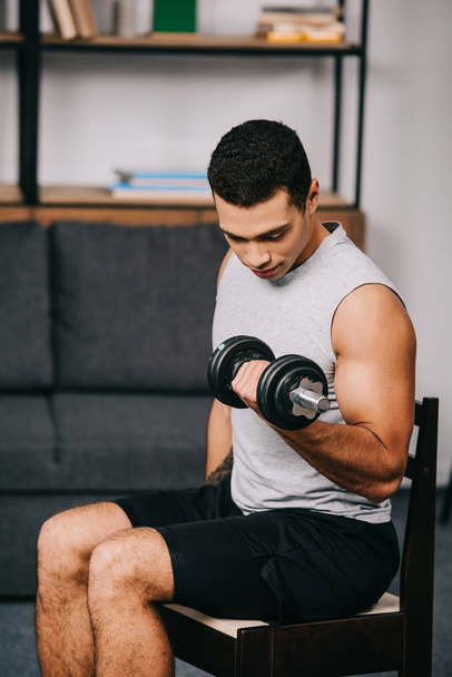 strong mixed race man training in sportswear with heavy dumbbell on chair - Photo, Image