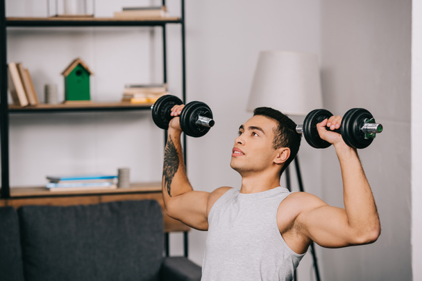 strong mixed race sportsman holding heavy dumbbells and exercising in living room  - Φωτογραφία, εικόνα