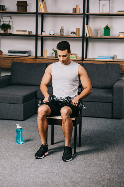 mixed race athlete sitting with dumbbells on chair in home gym - Fotoğraf, Görsel
