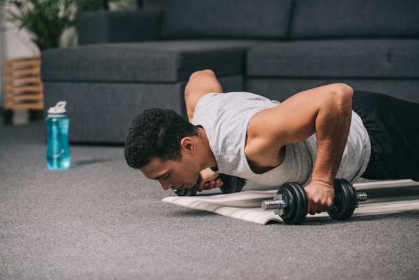 mixed race man doing push ups with dumbbells on fitness mat in living room  - Photo, Image