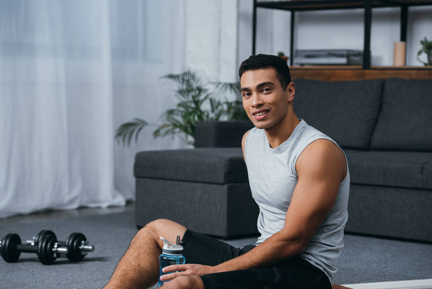 happy bi-racial man sitting with sport bottle in home gym  - Photo, Image