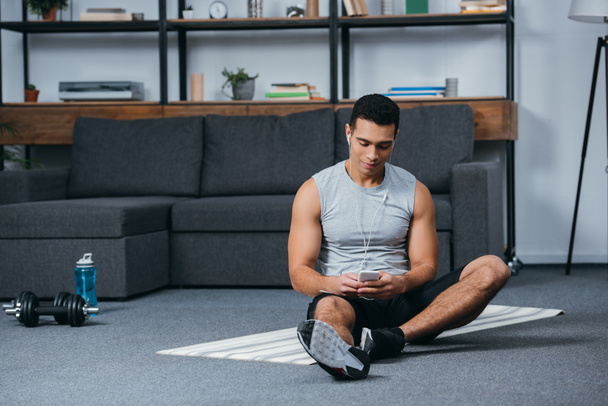 hombre bi-racial sosteniendo el teléfono inteligente y escuchar música en los auriculares en la alfombra de fitness
 - Foto, Imagen