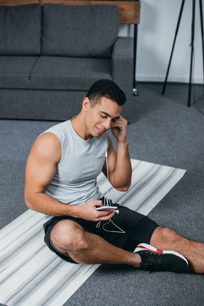 happy bi-racial man putting earphones in ear and holding smartphone while sitting on  fitness mat - 写真・画像