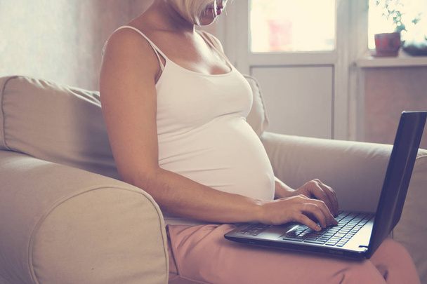 Young beautiful pregnant woman working on laptop. Pregnant business woman searching information and online shopping on laptop prepare for childbirth and parenting on sofa at home - 写真・画像