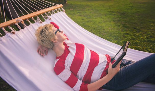 jonge vrouw met behulp van een tablet-pc terwijl u ontspant op hangmat in een rustige tuin tijdens vakantie - Foto, afbeelding