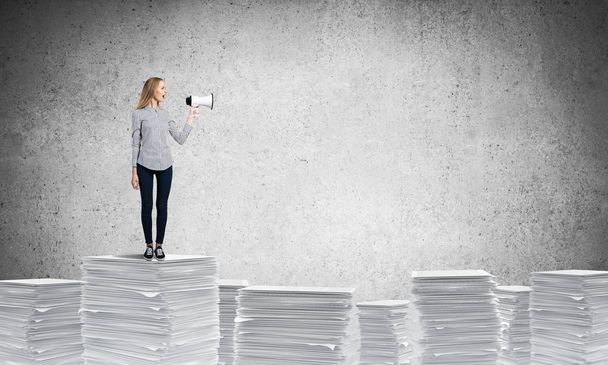 Kobieta w odzieży casual, stojąc na stos dokumentów z głośnika w parze z szarej ścianie na tle. Techniki mieszane. - Zdjęcie, obraz