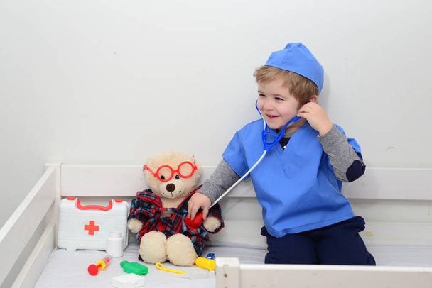 Bambino giocare medico o infermiere con peluche orso giocattolo a casa. Il ragazzo felice ascolta uno stetoscopio per giocare. Bambino giocherellone che gioca. Adorabile bambino vestito da medico che gioca con il giocattolo su sfondo bianco
 - Foto, immagini