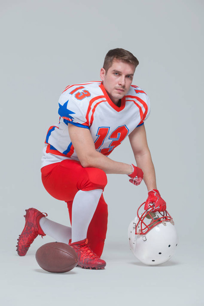 Американский футболист сидит на коленях с мячом и шлемом на сером фоне
 - Фото, изображение